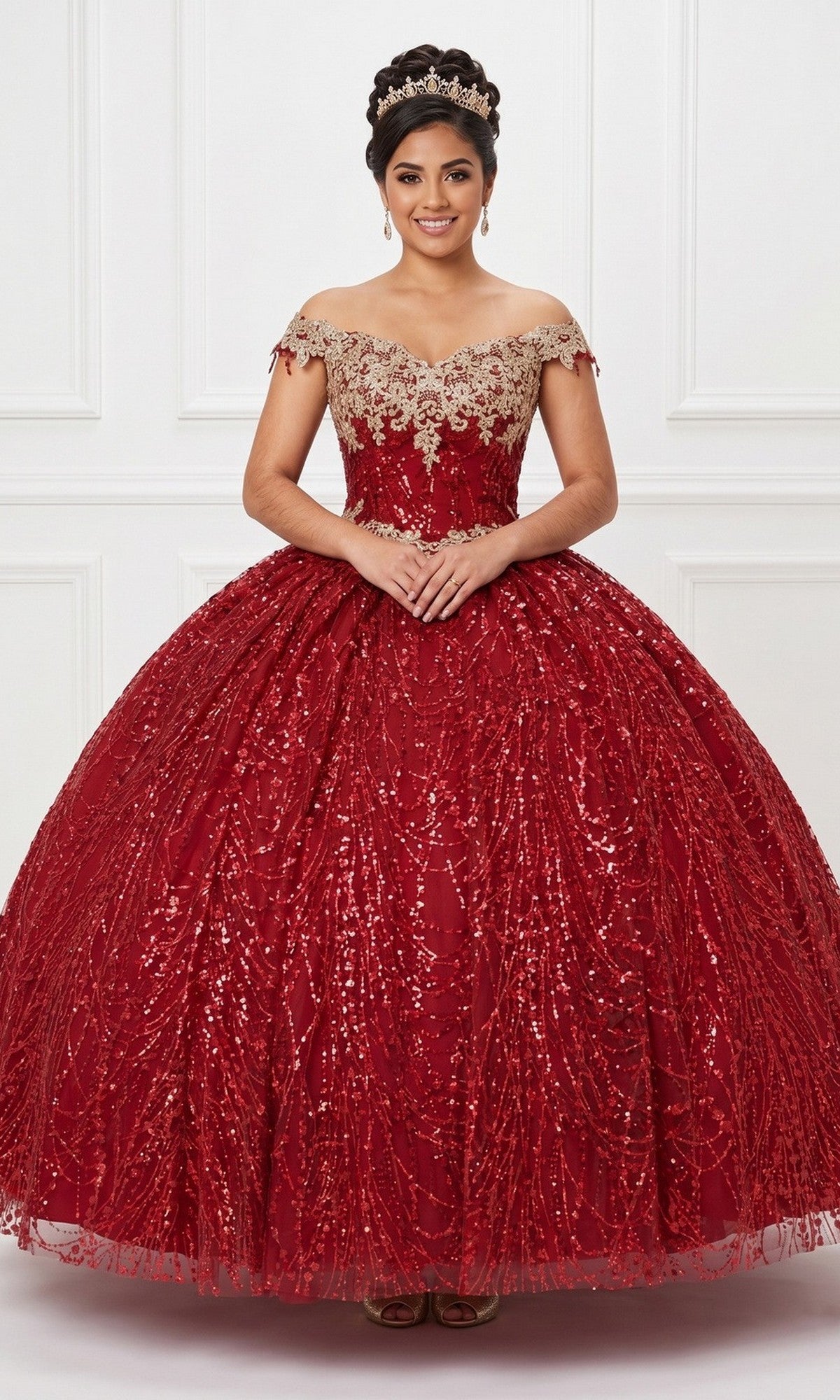 A young woman wearing the Quinceanera Dress CHF3011 By Chicas in burgundy, featuring an ornate off-the-shoulder design and corset back, with a tiara on her head, stands with hands clasped in front of a white decorative wall.